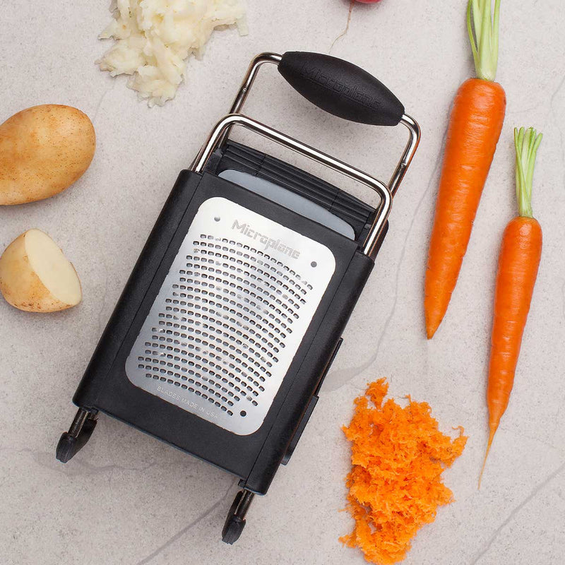 Microplane | 4-Sided Professional Box Grater on countertop with whole and grated carrots and potato; features ergonomic handle, fold-out feet, and versatile blade styles for precise grating.