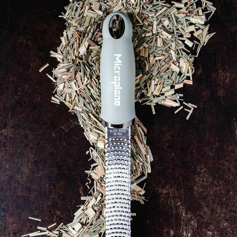 Microplane | Premium Zester, Grater - Classic Blue with finely-toothed stainless-steel blade on dark cutting surface, surrounded by shredded lemongrass, showcasing its precision and versatility.