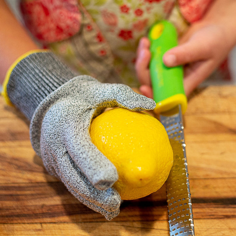 Child using a Microplane Cut Resistant Kitchen Safety Glove while zesting a lemon with a handheld grater on a cutting board.