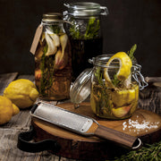 Microplane | Master Series Zester with Walnut Handle on a rustic kitchen slab, surrounded by citrus, herbs, and infused jars, perfect for precise zesting and grating.