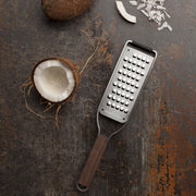 Microplane Master Series Extra Coarse Grater with Walnut Handle sits beside a halved coconut and shredded coconut pieces, showcasing its precision and ergonomic design for versatile kitchen use.