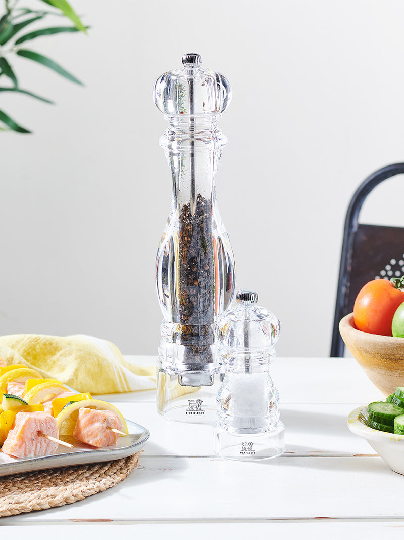 Peugeot Paris Nancy Pepper Mill in acrylic, 9 inches, next to a salt grinder, amid salmon skewers and fresh produce on a kitchen table.