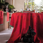Le Jacquard Francais | Féerie Tablecloth, Red, 69 x 98, drapes elegantly over a festive dining table, accentuating a cozy holiday setting with holiday decor and a hidden Scottish Terrier underneath.