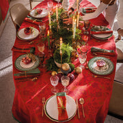 Féerie Tablecloth in red enhances a festive tablescape with elegant gold flatware, evergreen centerpiece, and warm candlelight, embodying traditional Christmas charm.