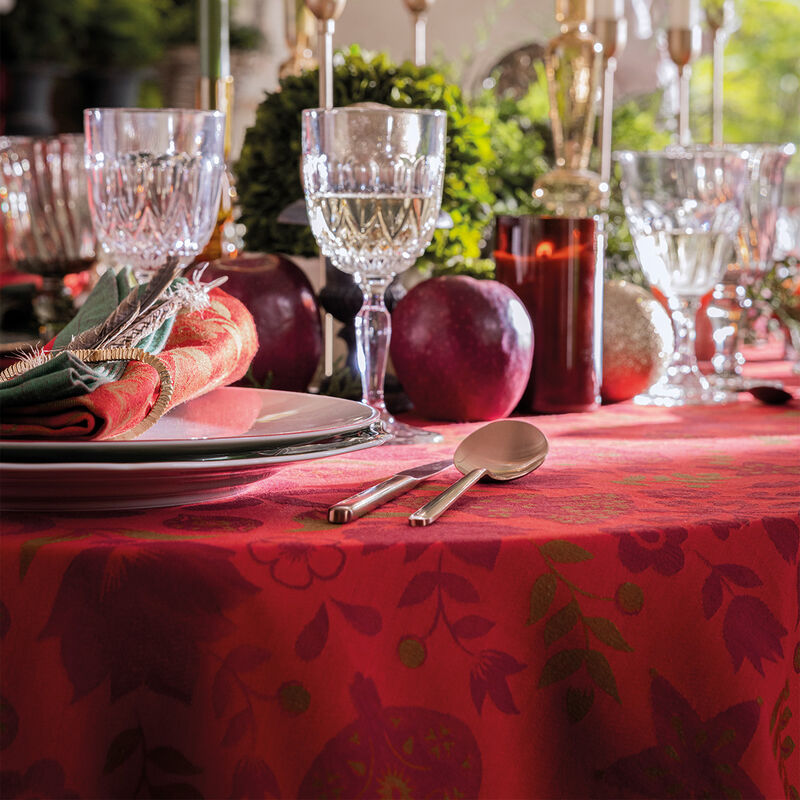 Le Jacquard Francais Féerie Tablecloth in red showcases a festive table setting with patterned cloth, elegant crystal glassware, and greenery, evoking a folkloric Christmas ambiance.
