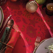 Festive table setting with Le Jacquard Francais Féerie Tablecloth in red, featuring gold floral motifs, complemented by elegant dinnerware, a lit candle, and decorative accents.