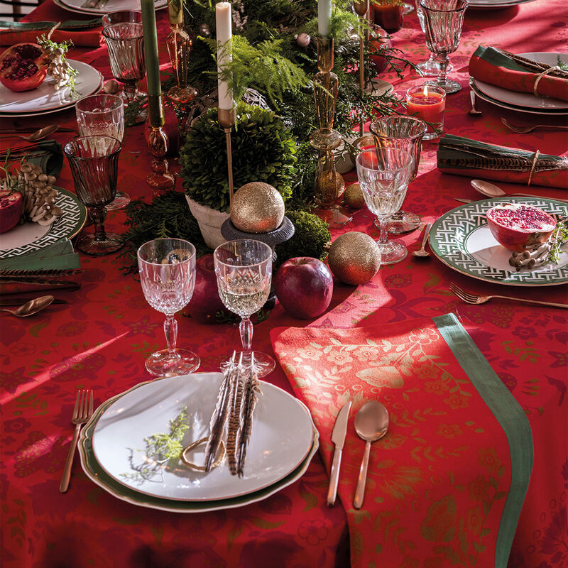Le Jacquard Francais Féerie Tablecloth in red elegantly sets a festive table with gold flatware, crystal goblets, and a lush evergreen centerpiece.