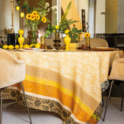 Le Jacquard Francais Gipsy Tablecloth in yellow adorns a dining table with floral motifs, amber glassware, daisies, sunflowers, and fresh lemons.