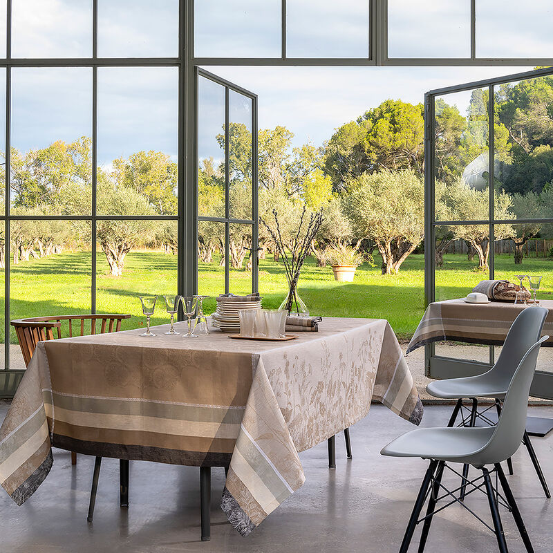 Dining space showcasing Le Jacquard Francais Bucolique Tablecloth, Beige, on tables set with glassware and vases, overlooking sunlit olive grove through open French doors.