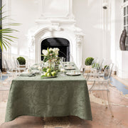 Le Jacquard Francais Tivoli Tablecloth in Green elegantly drapes a formal dining setup with china and floral centerpiece, surrounded by clear ghost chairs in a refined dining room.
