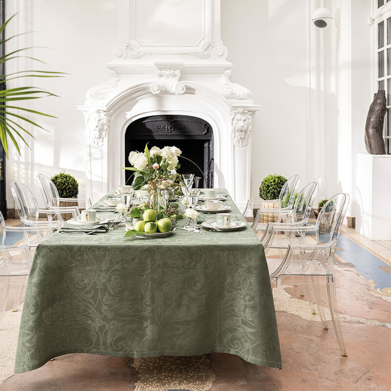 Le Jacquard Francais Tivoli Tablecloth in Green elegantly drapes a formal dining setup with china and floral centerpiece, surrounded by clear ghost chairs in a refined dining room.
