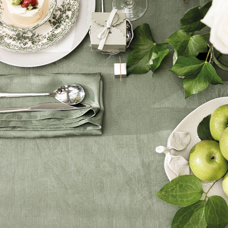 Le Jacquard Francais | Tivoli Tablecloth, Green, 69 x 98, elegantly drapes a table adorned with a pastry plate, cutlery, green apples, and a ribbon-tied gift box.