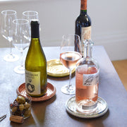Sir|Madam Brass Bottle Coaster elegantly holds a rosé bottle on a silver saucer, amidst a casual wine-and-snacks setup, showcasing its vintage-inspired charm.