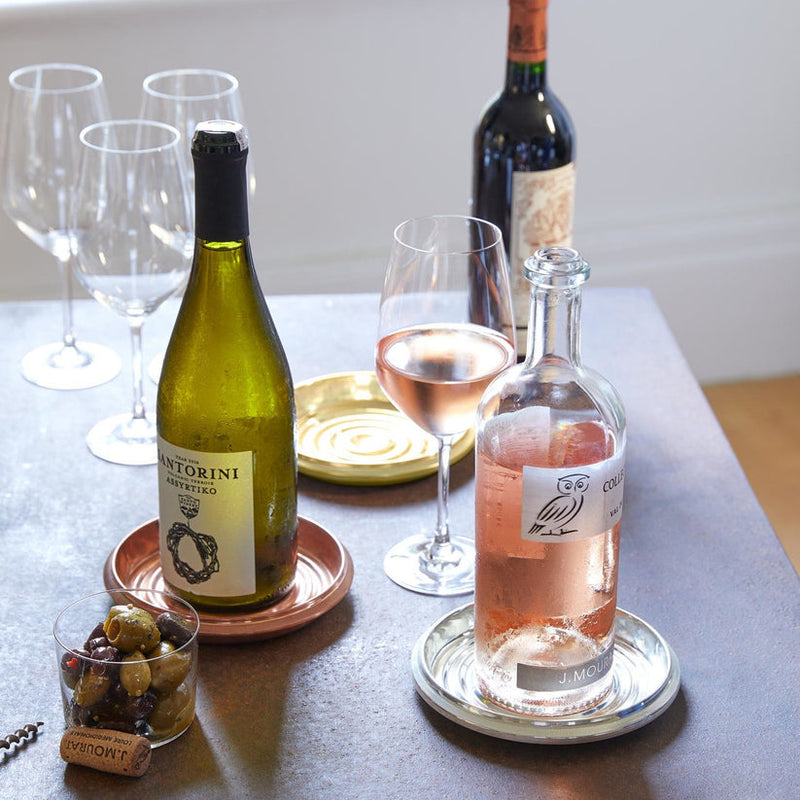 Sir|Madam Brass Bottle Coaster elegantly holds a rosé bottle on a silver saucer, amidst a casual wine-and-snacks setup, showcasing its vintage-inspired charm.