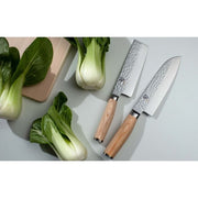 Shun Premier Blonde Santoku 7 knife with hammered finish and light handle, displayed on a wooden board with four baby bok choy and another Japanese knife.