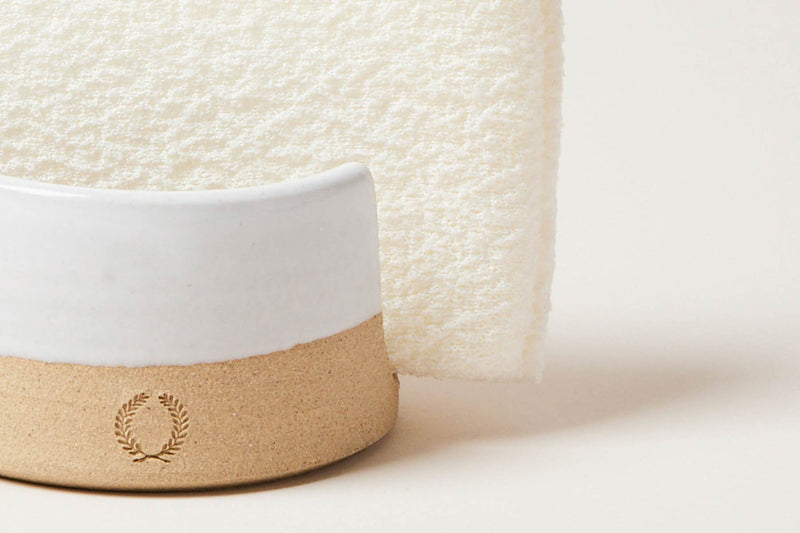 Farmhouse Pottery Sponge Holder cradles a folded textured cloth, showcasing a two-tone ceramic design with a laurel-wreath motif, perfect for enhancing kitchen organization.