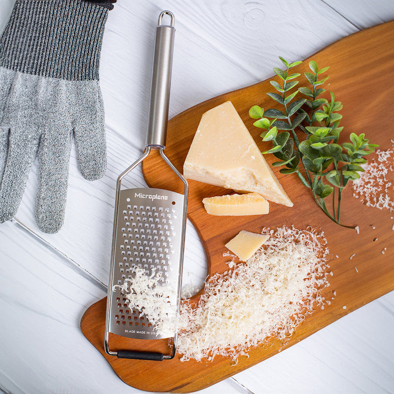 Microplane Professional Series Coarse Grater on a wooden board with grated cheese, protective glove, and cheese wedge; showcases precision grating for culinary excellence.