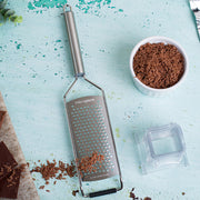 Microplane Professional Series Coarse Grater setup for chocolate grating, featuring a stainless steel design, ergonomic handle, and clear catch container alongside chocolate shavings and pieces.