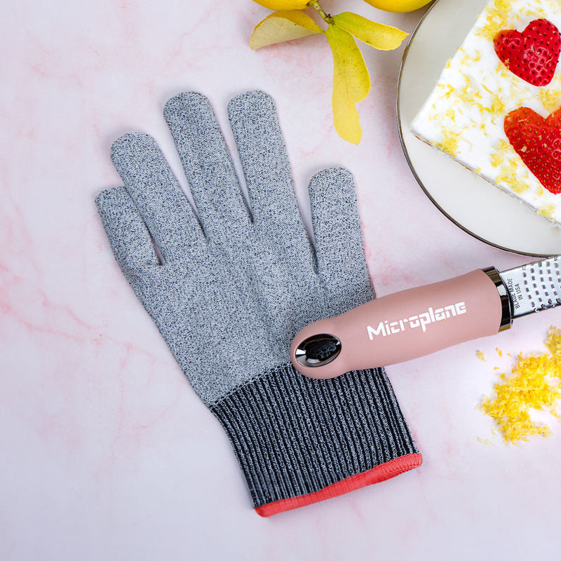 Microplane | Cut Resistant Kitchen Safety Gloves alongside a pink-handled Microplane grater, lemons, and a frosted cake slice, on a marble-style surface.