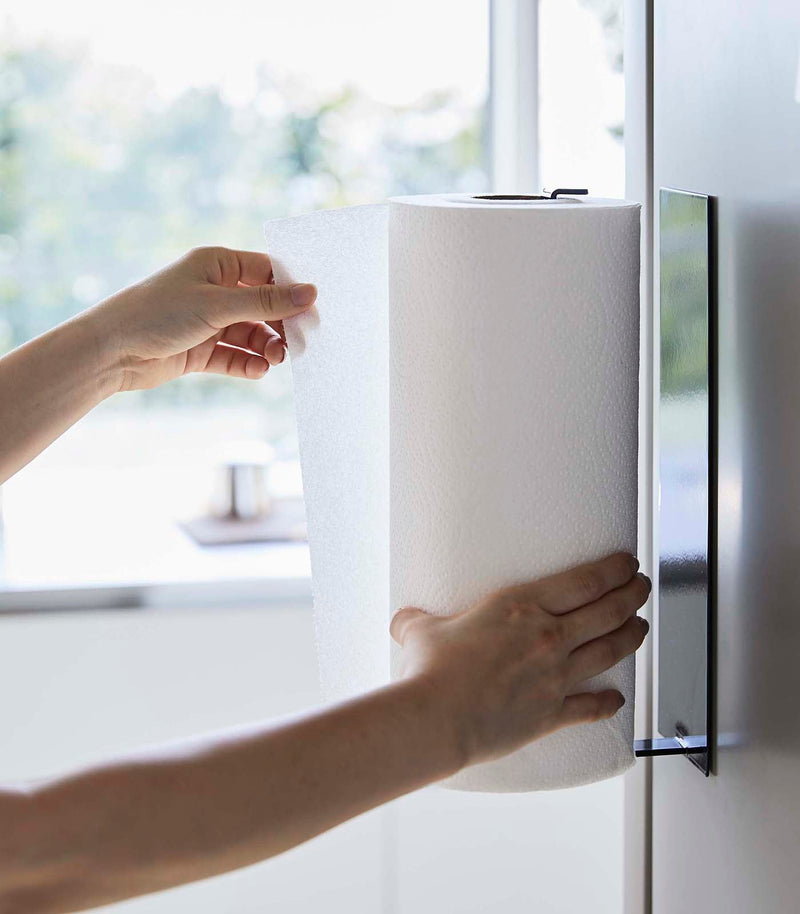 Yamazaki Home | Magnetic Steel Paper Towel Holder, White, mounted on a fridge, with hands pulling a sheet, showcasing its space-saving design in a modern kitchen.