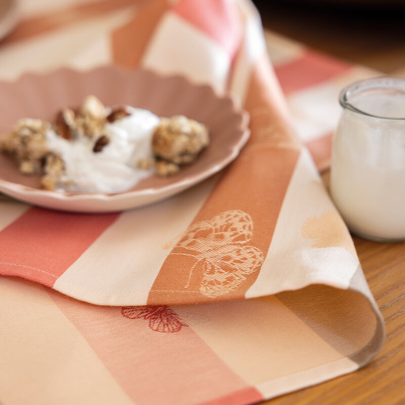 Le Jacquard Francais | Bal Des Papillons Napkin in pink, featuring butterfly motifs, under a ceramic plate with granola and yogurt on a wooden surface.