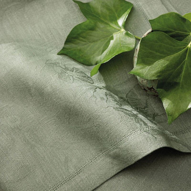 Le Jacquard Francais | Tivoli Napkin, Green: Close-up of a soft, muted-green linen napkin with a woven border, adorned with glossy ivy leaves.