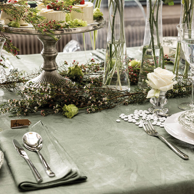 Elegant tablescape showcasing a Le Jacquard Francais Tivoli Napkin in green, paired with silverware, mini cakes, and floral accents for a sophisticated dining experience.