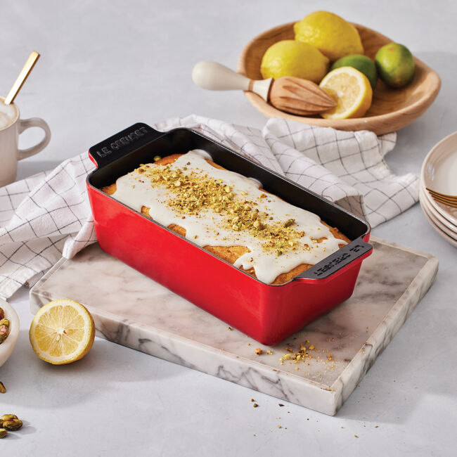 Le Creuset | Signature Loaf Pan - Sea Salt with lemon loaf cake topped with glaze and pistachios, surrounded by lemons, pistachios, and baking tools on a marble board.