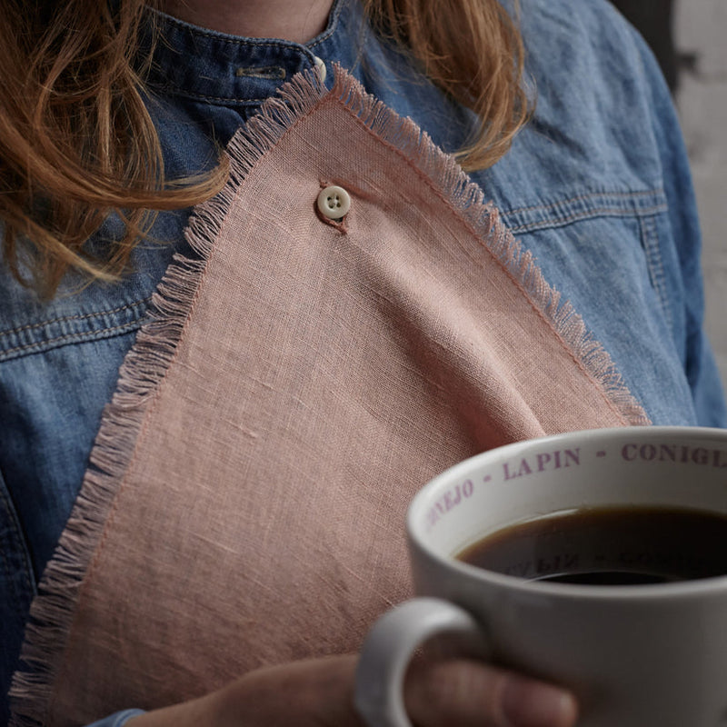 Person holds a large mug of coffee, wearing a blue shirt with a Sir|Madam Linen Frayed Edge Napkin draped over their chest, buttoned at the collar.