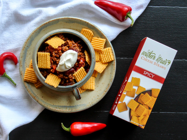 Southern Straws | Cheese Straws accompany a hearty chili bowl, showcasing the Spicy variety’s packaging, surrounded by fresh chili peppers and a rustic dining setup.