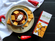 Southern Straws | Cheese Straws beside a chili con carne mug, topped with crackers, showcasing hand-crafted, spicy cheese wafers from a family recipe, ideal for snacking.