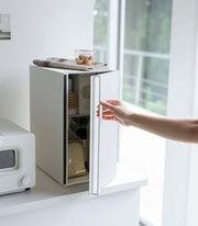 Yamazaki Home Bread Box showcased in a minimalist kitchen, emphasizing its vertical steel design for keeping baked goods fresh, with a fridge and toaster-oven nearby.