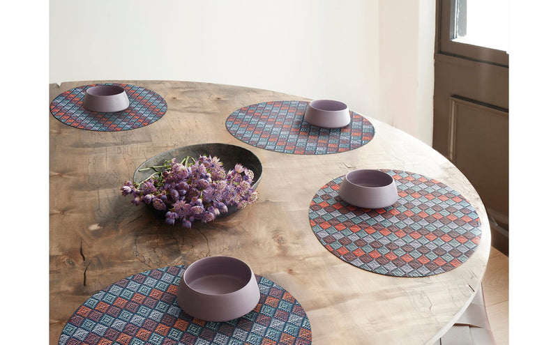 Chilewich | Kite Placemats - Gemstone - Round on a table, featuring a vibrant geometric design with a bowl of purple flowers, enhancing tableware aesthetics.