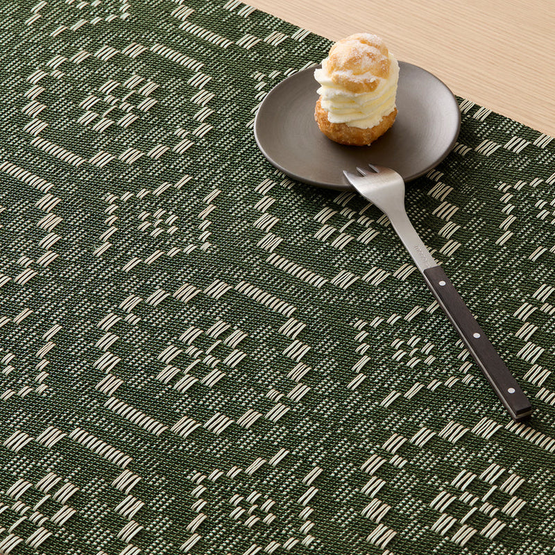 Chilewich | Overshot Rectangular Placemat, Thyme, showcases interlocking circles under a cream-puff pastry on a side plate, complemented by a fork/knife with a wooden handle.