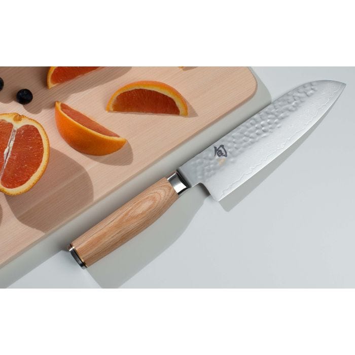 Shun Premier Blonde Santoku, 7, set on a kitchen counter with citrus wedges and blueberries on a wooden board, showcasing its hammered finish and light wood handle.