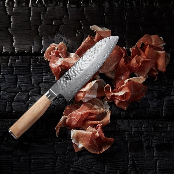 Shun Premier Blonde Santoku, 7, with a hammered finish and light wooden handle, displayed on sliced cured ham on a charred wood surface.