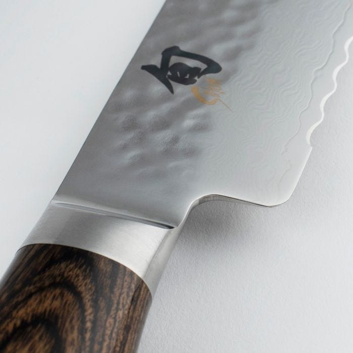 Shun | Premier Bread Walnut, 9: Close-up of a santoku-style blade with tsuchime texture, Damascus grain, Japanese logo, and polished bolster with dark patterned wooden handle.
