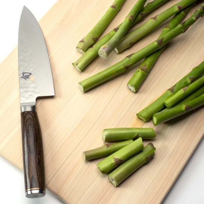 Shun Premier 6-inch Chef's Knife with a hammered steel blade and dark wooden handle, displayed beside trimmed asparagus on a light-wood cutting board.
