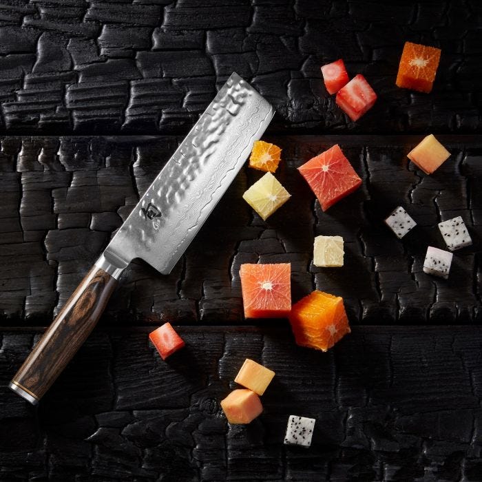 Overhead view of Shun Premier Nakiri Walnut, 5.5, amidst diced fruit, showcasing its hammered finish blade and polished wood handle on a dark wooden surface.