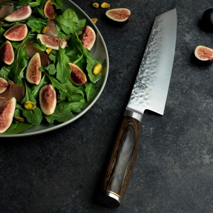 Shun Premier Kiritsuke Walnut, 8, with a hammered stainless blade and dark wooden handle, lies beside a fig and arugula salad on a countertop.