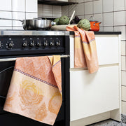 Le Jacquard Francais | Jardin Des Choux Tea Towel in orange, draped over a black stove, with artichokes and cookware on the countertop.