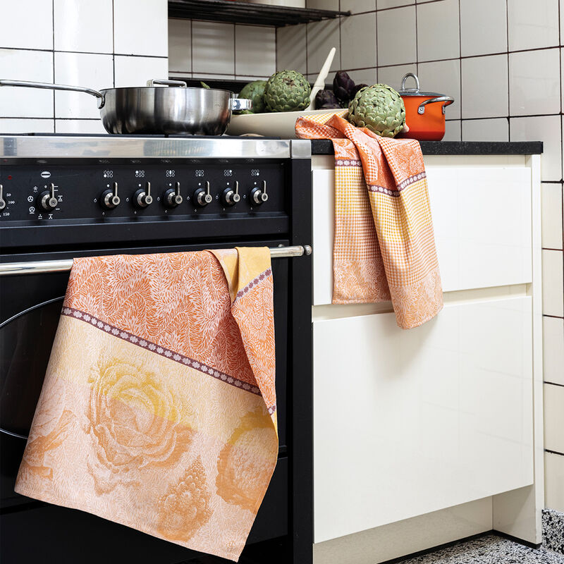Le Jacquard Francais | Jardin Des Choux Tea Towel in orange, draped over a black stove, with artichokes and cookware on the countertop.