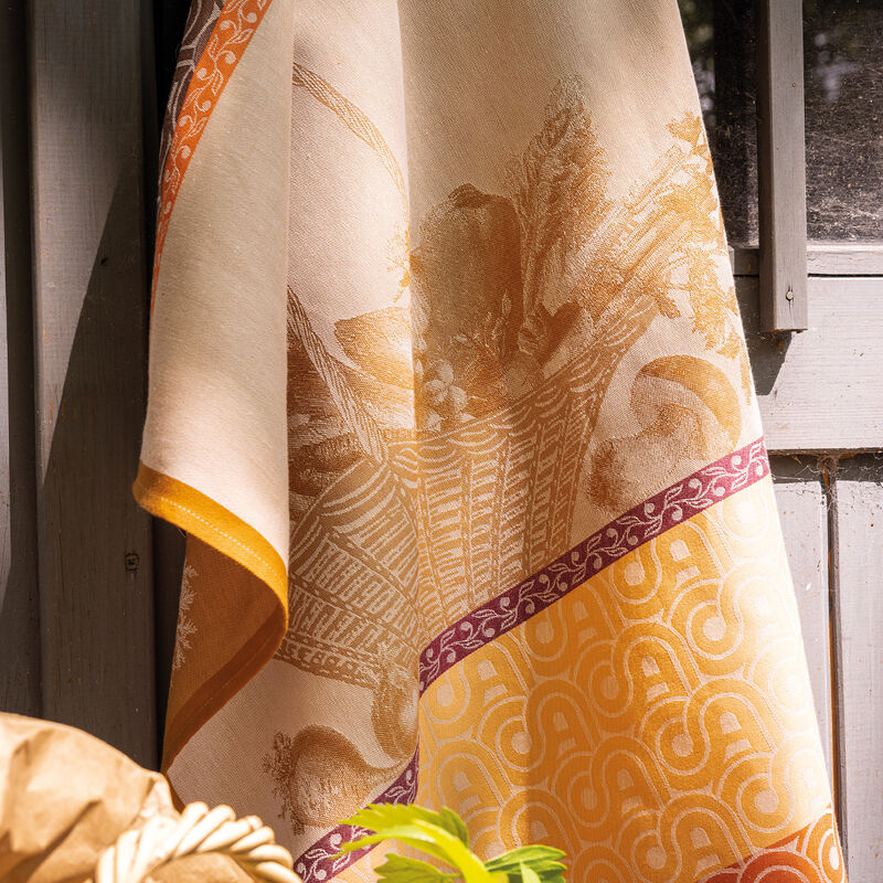 Le Jacquard Francais | Potager D'Hiver Tea Towel, Orange, featuring a woven cornucopia motif, draped over a wooden surface with a paper bag in view.