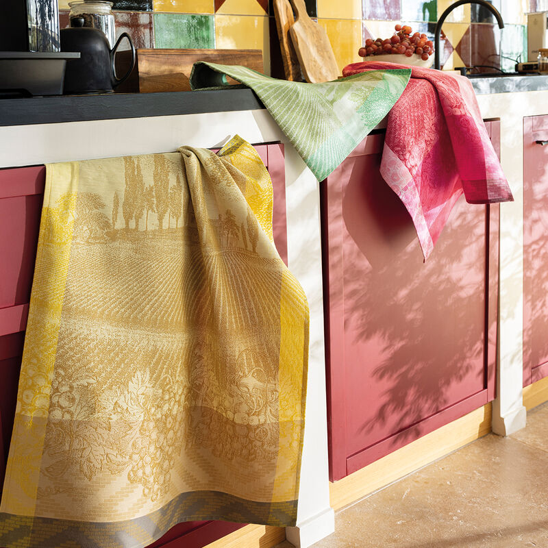 Le Jacquard Français | Vignoble Tea Towel, Yellow, draped on red cabinets in a cozy kitchen, featuring vineyard design, with cutting board and teapot on the countertop.