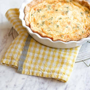 Golden-brown quiche in a fluted white ceramic pan atop a Caravan Home 20x30 Gingham Dijon & Grey Towel on a marble countertop.