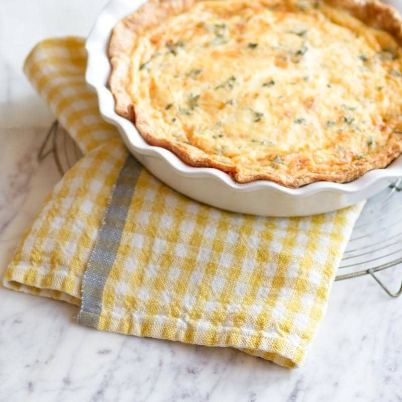 Golden-brown quiche in a fluted white ceramic pan atop a Caravan Home 20x30 Gingham Dijon & Grey Towel on a marble countertop.