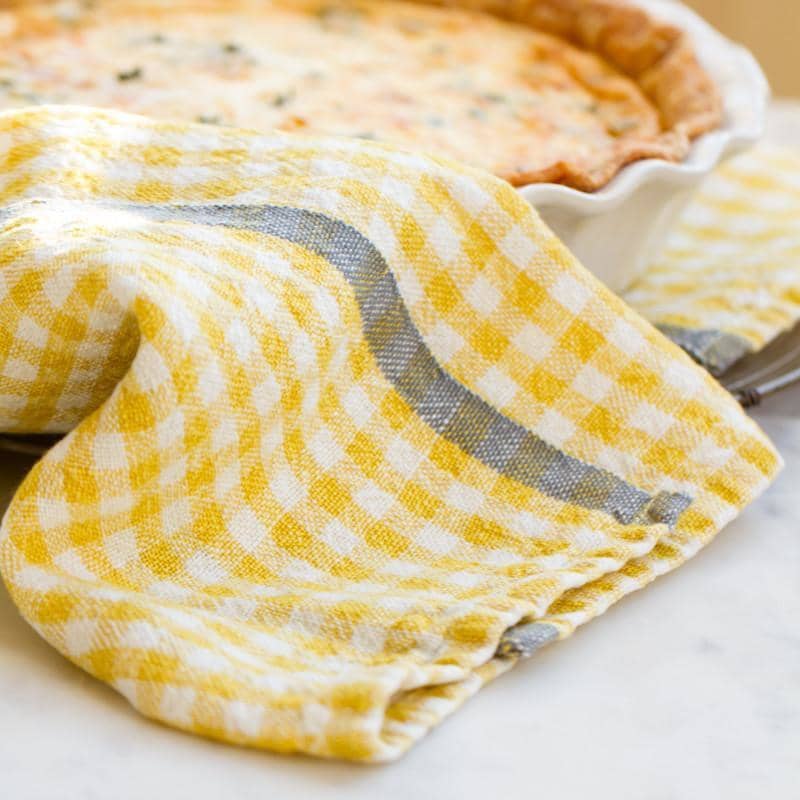 Caravan Home's 20 x 30 Gingham Dijon & Grey Towel draped over a ceramic dish with a pie, on a marble countertop.