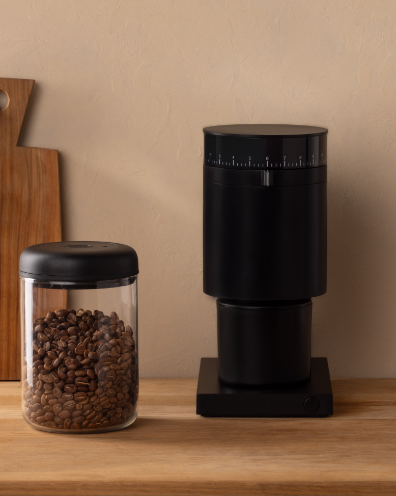 FELLOW | Opus Conical Burr Grinder, Matte Black, with grind-size adjustment, sits on a countertop alongside a jar of coffee beans and a cutting board.