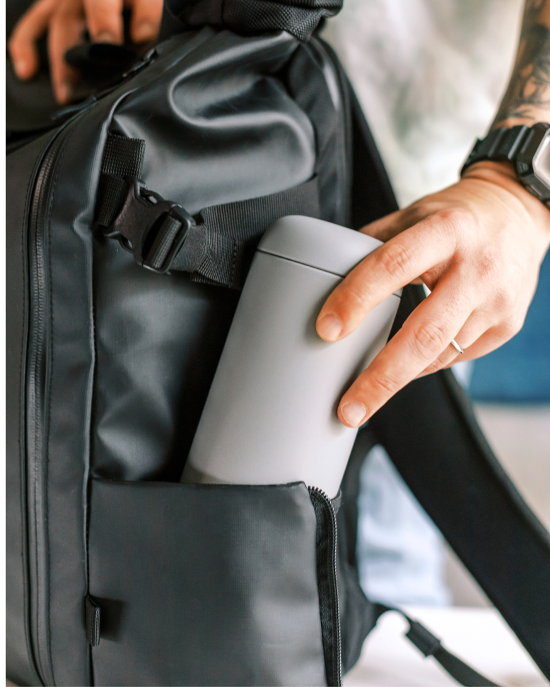 Hand places a matte-gray FELLOW - Carter Move Mug into a black backpack's side pocket, showcasing its leak-proof, travel-friendly design.