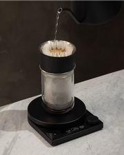 FELLOW | Tally Pro Precision Scale Studio Edition, Black, displayed mid-brew, enhancing pour-over coffee precision with its OLED display and Brew Assist™ Mode on a marble countertop.